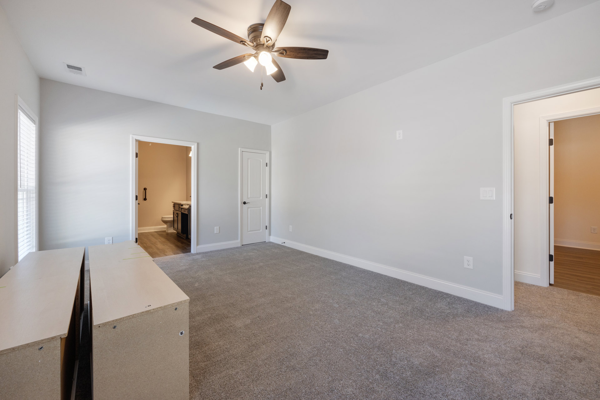 Ceiling fan with light fixture mounted on white plaster ceiling, wooden box on light-colored flooring, white door with black handle, partial view of bathroom with toilet and sink