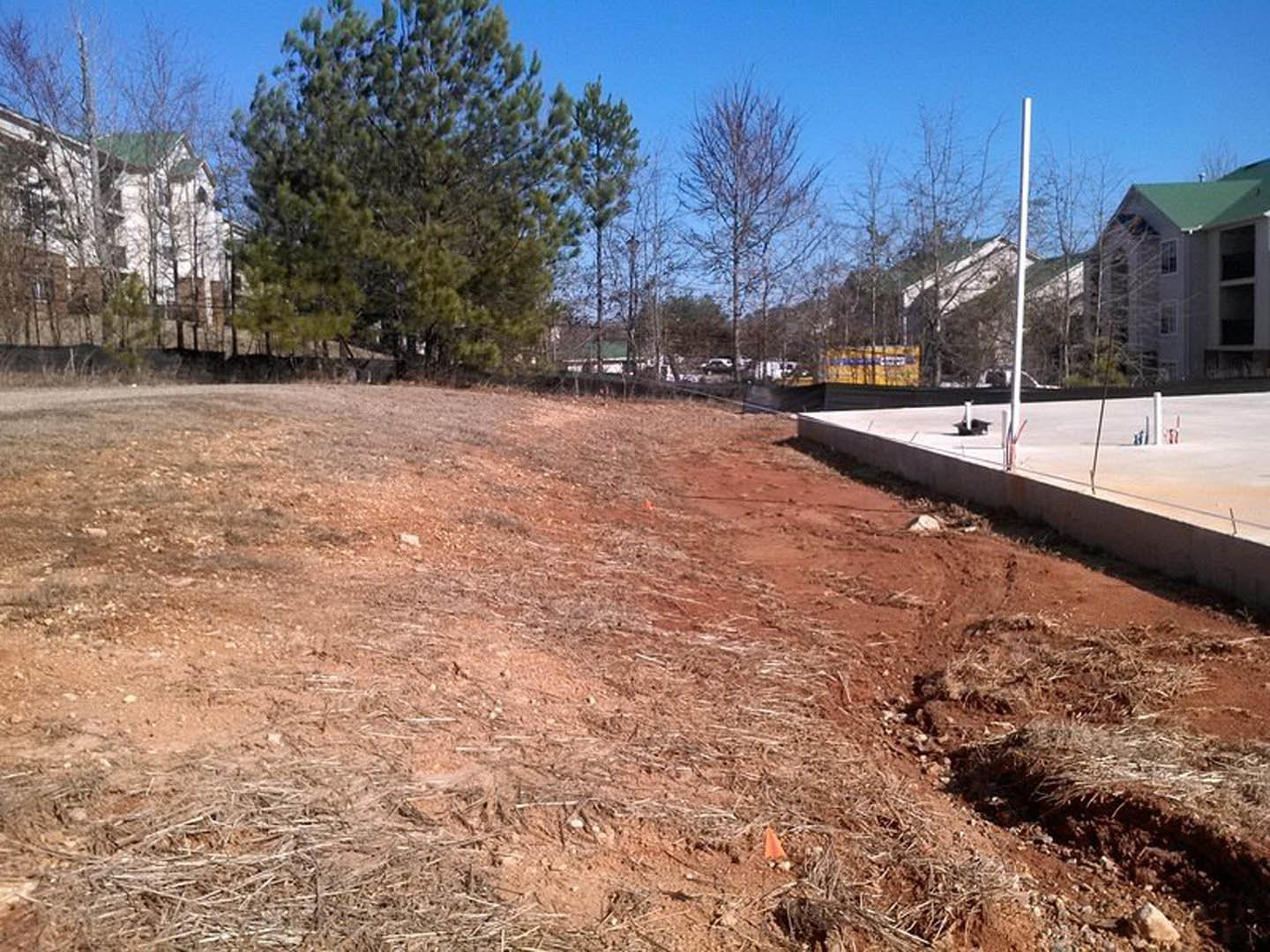 Dirt road bordered by leafy trees and a wooden fence, custom home with green roof partially visible in background