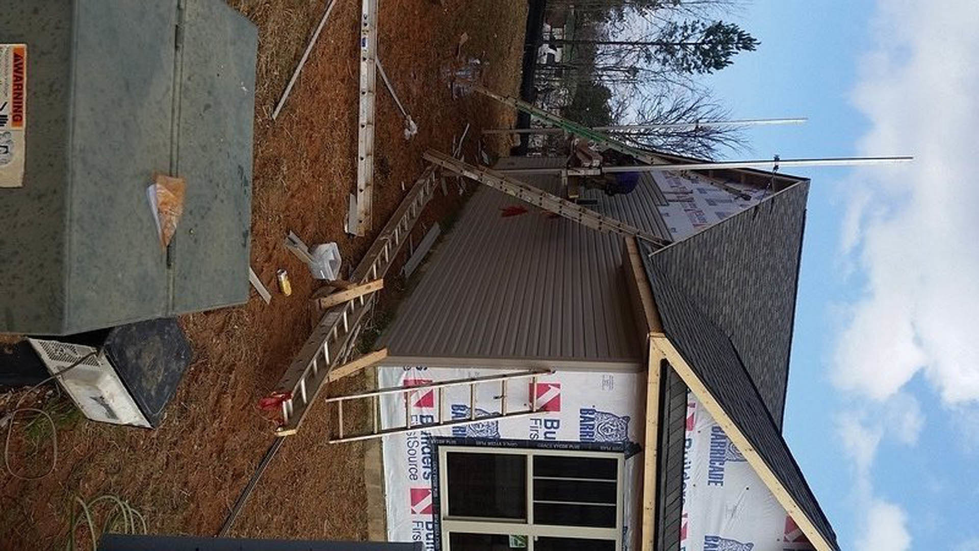 Aluminum ladder leaning against gray siding of two-story house, white-framed window above, cloudy sky in background, concrete walkway with scattered debris below