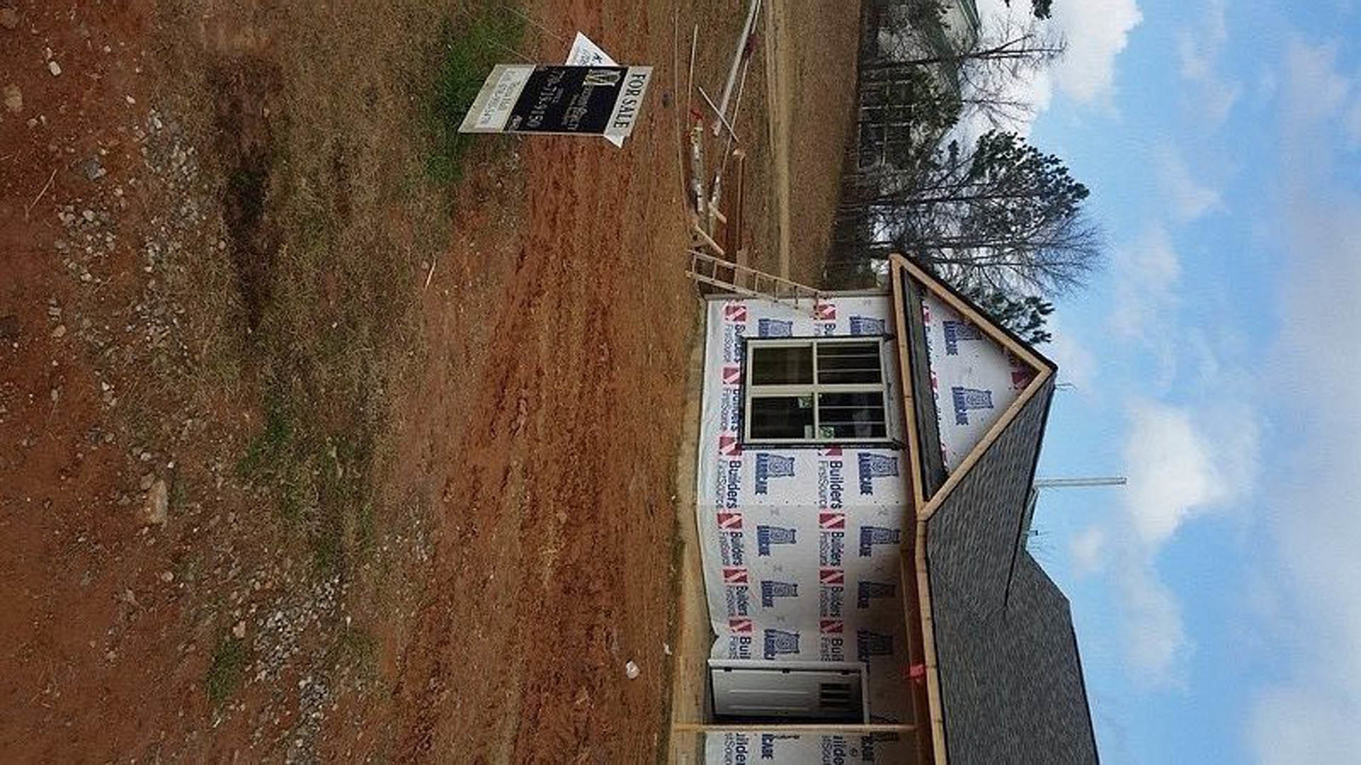 Partially finished house with gray siding, white trim, and exposed roof, set in a grassy yard under a cloudy sky