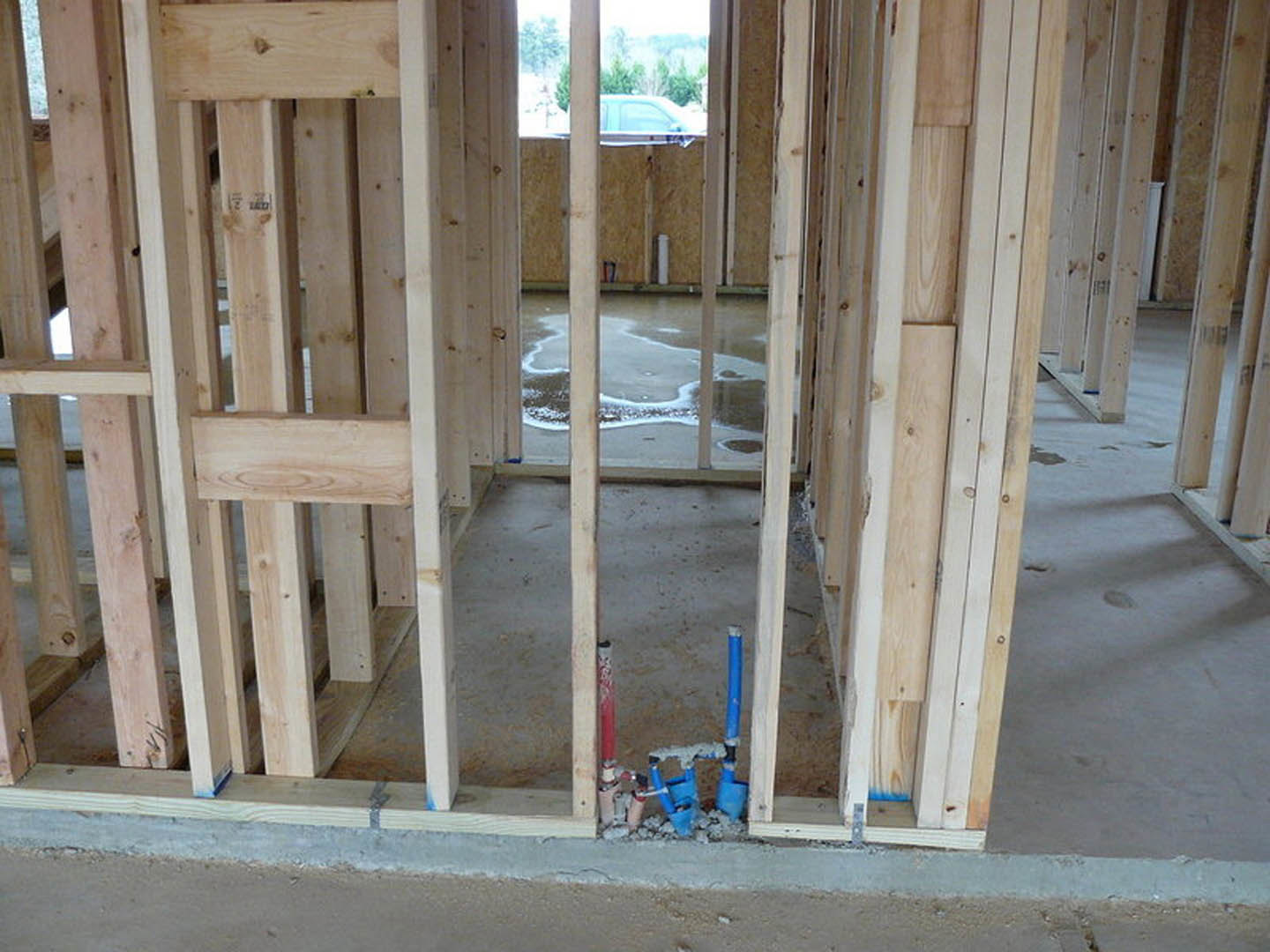 Wood framing and beams with exposed blue plumbing pipe, unfinished concrete wall, puddle on subfloor, window overlooking trees in background
