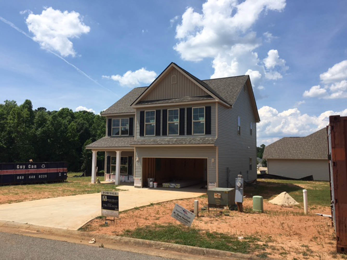 Modern two-story home with attached garage, white siding, large windows, covered front porch, landscaped yard with white stone accents, and mature trees in the background