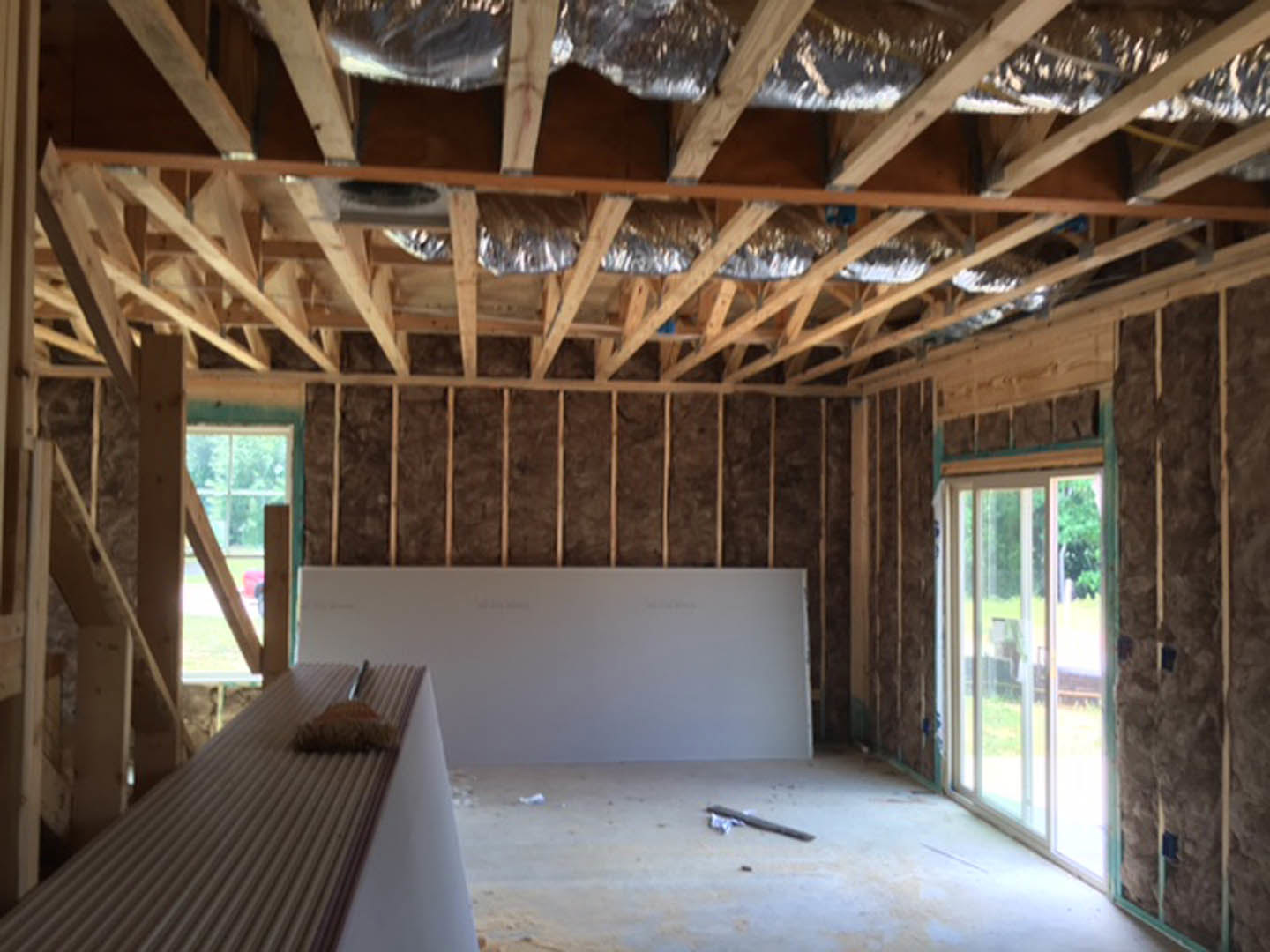 Room with exposed wood beams, silver foil insulation on ceiling, window with white trim, broom resting on shelf, whiteboard and pen nearby