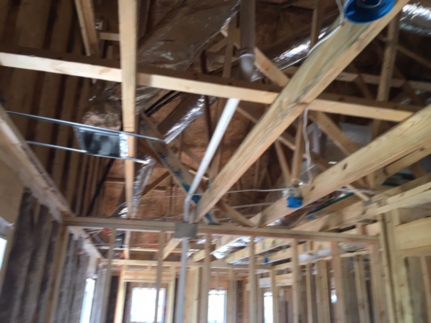 Exposed wooden beams and framing with visible pipes on unfinished house interior, blurred blue object and window in background