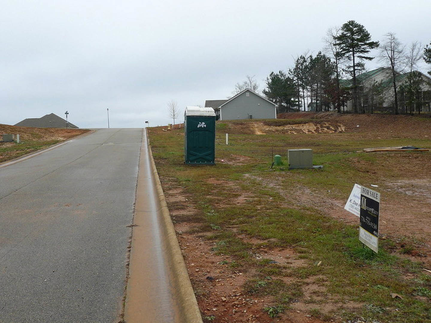 Grey house with white roof set back from asphalt road, green waste container with white logo near curb, black sign with white text on grassy verge, white utility box on ground