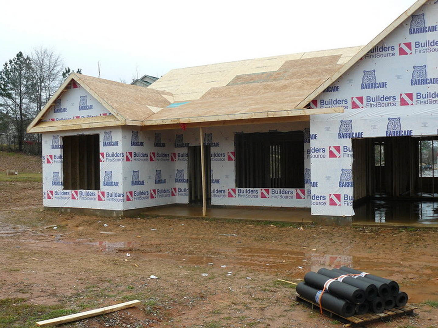 Partially built house with exposed wooden framing, rolls of black roofing material stacked near the foundation, window displaying a sign, red square with white zigzag pattern