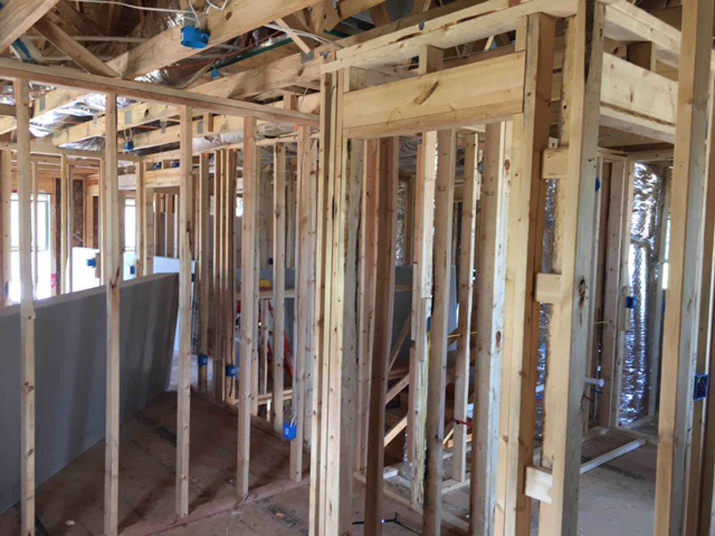 Exposed wooden framing and ceiling beams in a residential construction site, blue electrical outlet visible on ceiling, unfinished walls and insulation.
