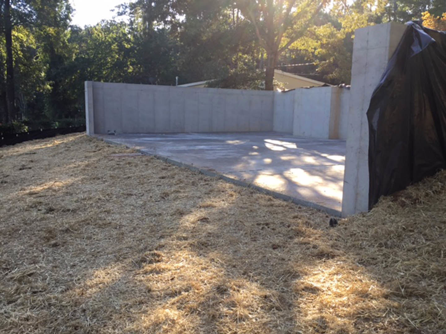 Concrete patio bordered by grass and trees, black bag resting on ground near textured concrete wall