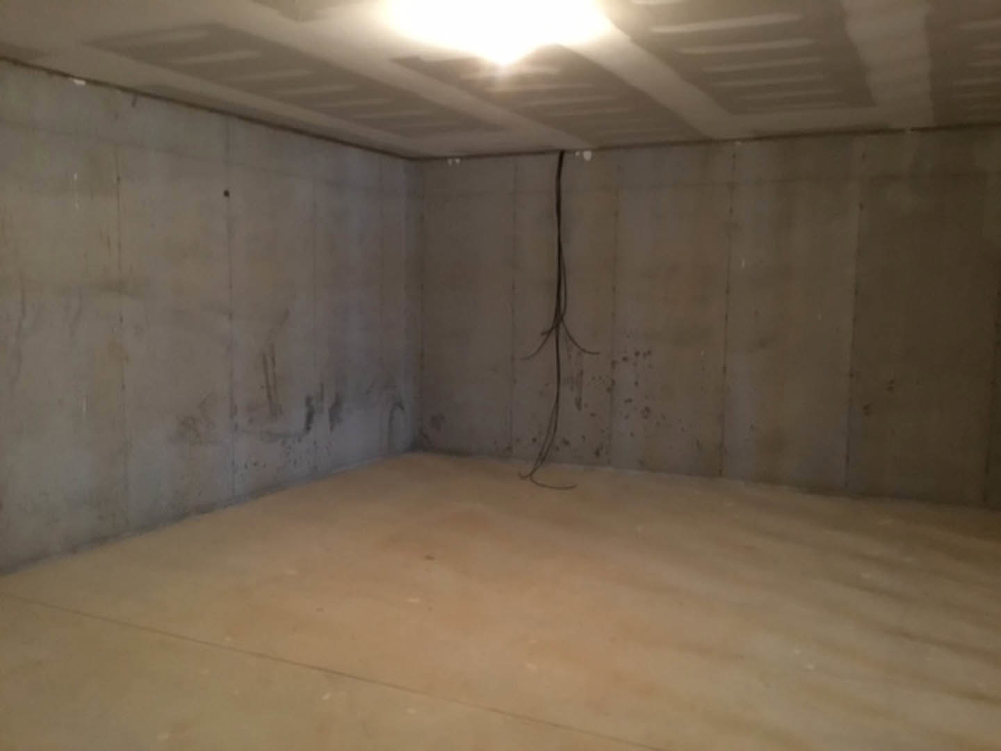 Concrete basement room with beige plaster walls, bright ceiling light, and black wire running along wall.