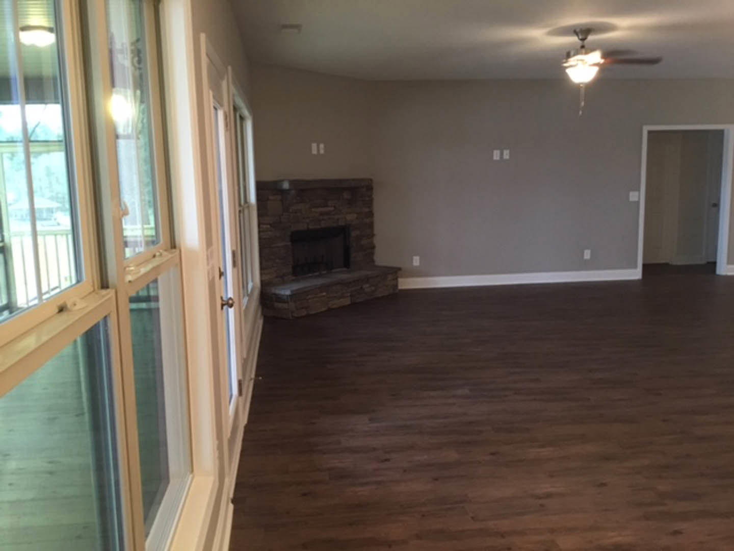 Living room with hardwood flooring, stone fireplace, ceiling fan, white door, and large window with dark trim