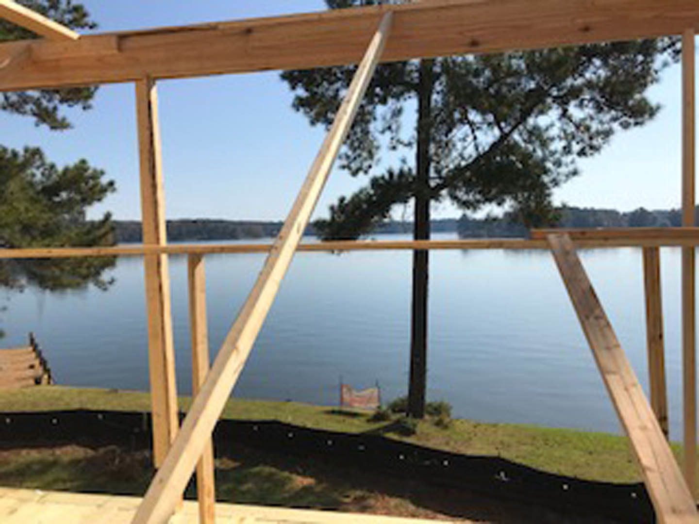 Wood-framed house under construction with exposed beams, surrounded by trees and a lake in the background