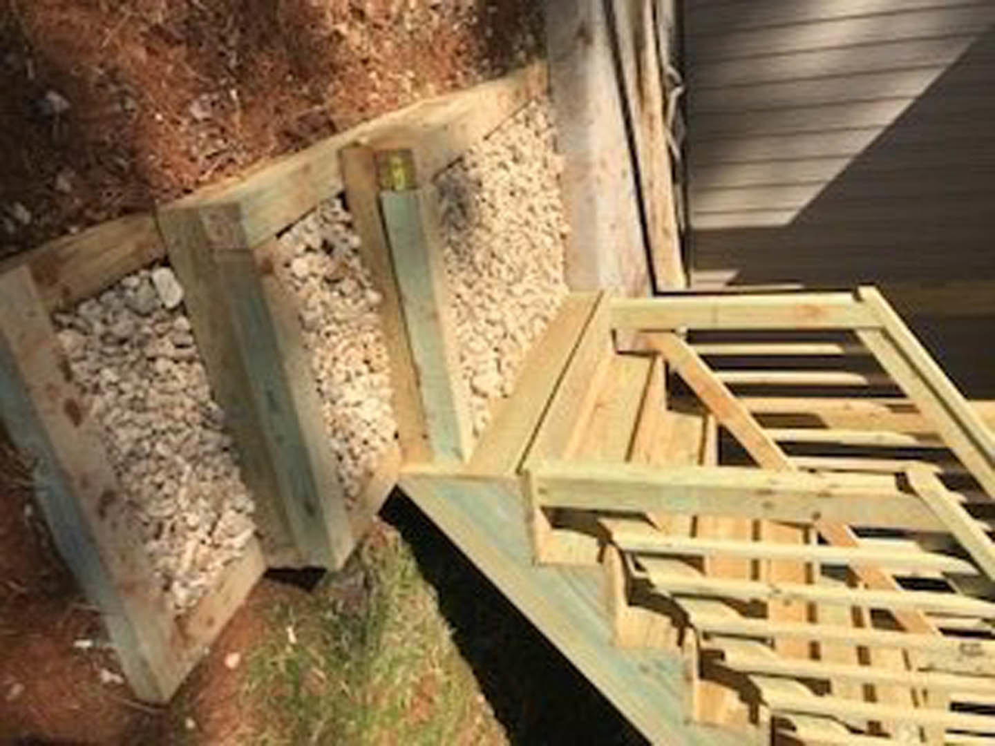 Wooden staircase with plank railing beside a pile of rocks contained in a wooden box, surrounded by grass and outdoor landscaping.