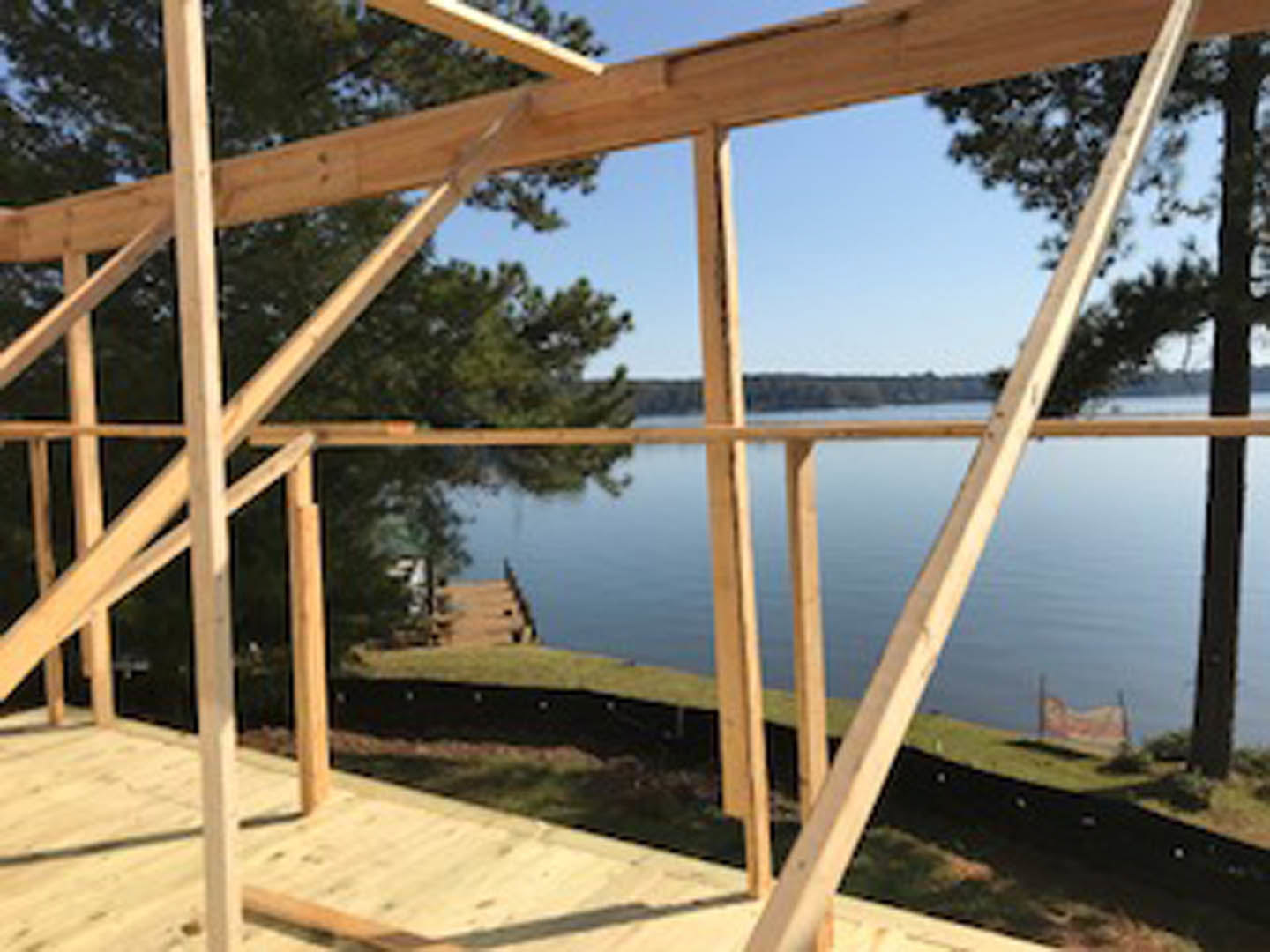 Wooden house frame with exposed beams and planks, set outdoors near trees and a lake under a clear sky