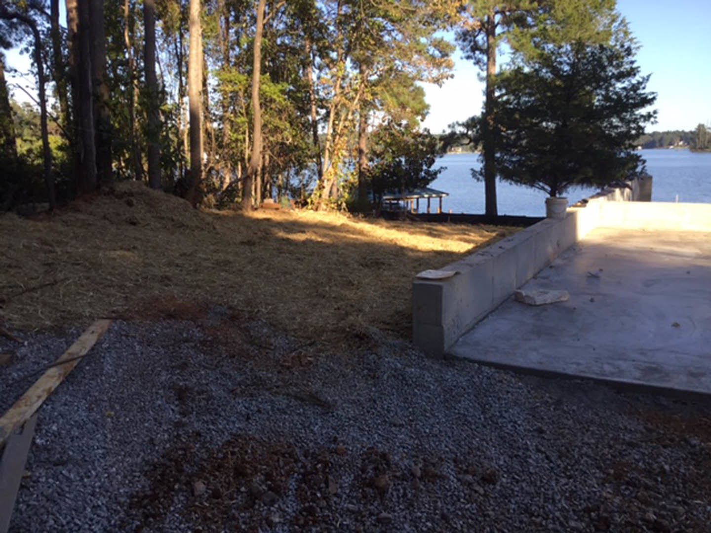 Concrete patio overlooking a lake, bordered by a tree and a low concrete wall with a white object placed on top; gravel and dirt pile visible near the patio, with a wooden board in