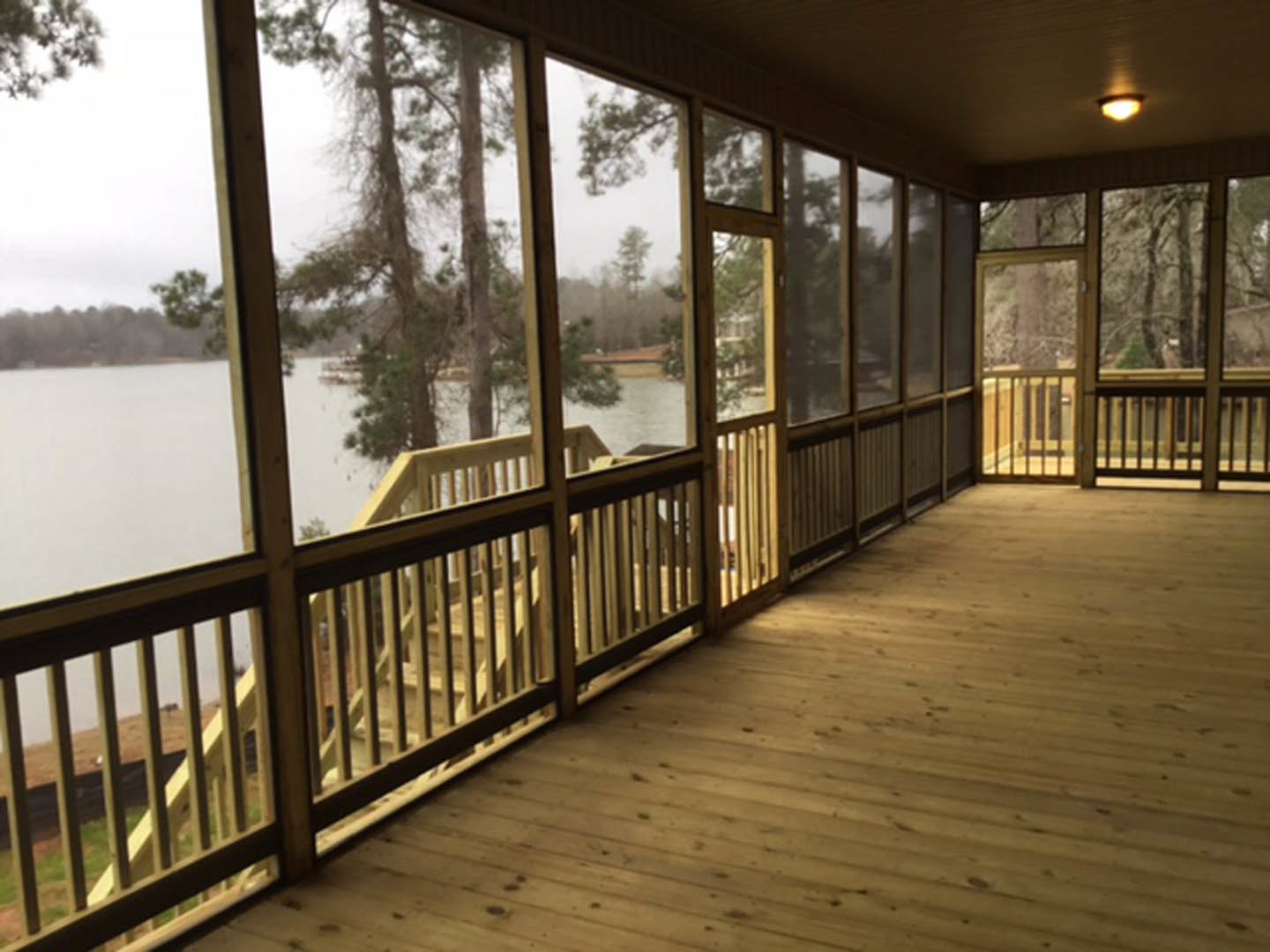 Wooden deck with metal railings overlooking a lake, screen door, ceiling light, large window framing trees, outdoor flooring