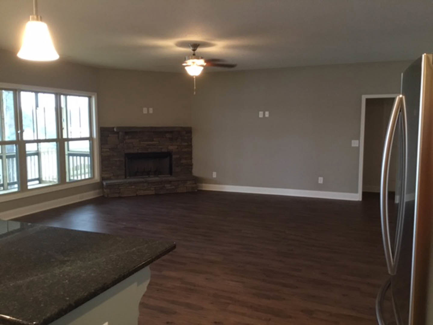 Living room featuring stone fireplace, ceiling fan, wood flooring, white cabinetry, black countertop, stainless steel refrigerator, large window with railing, and modern light