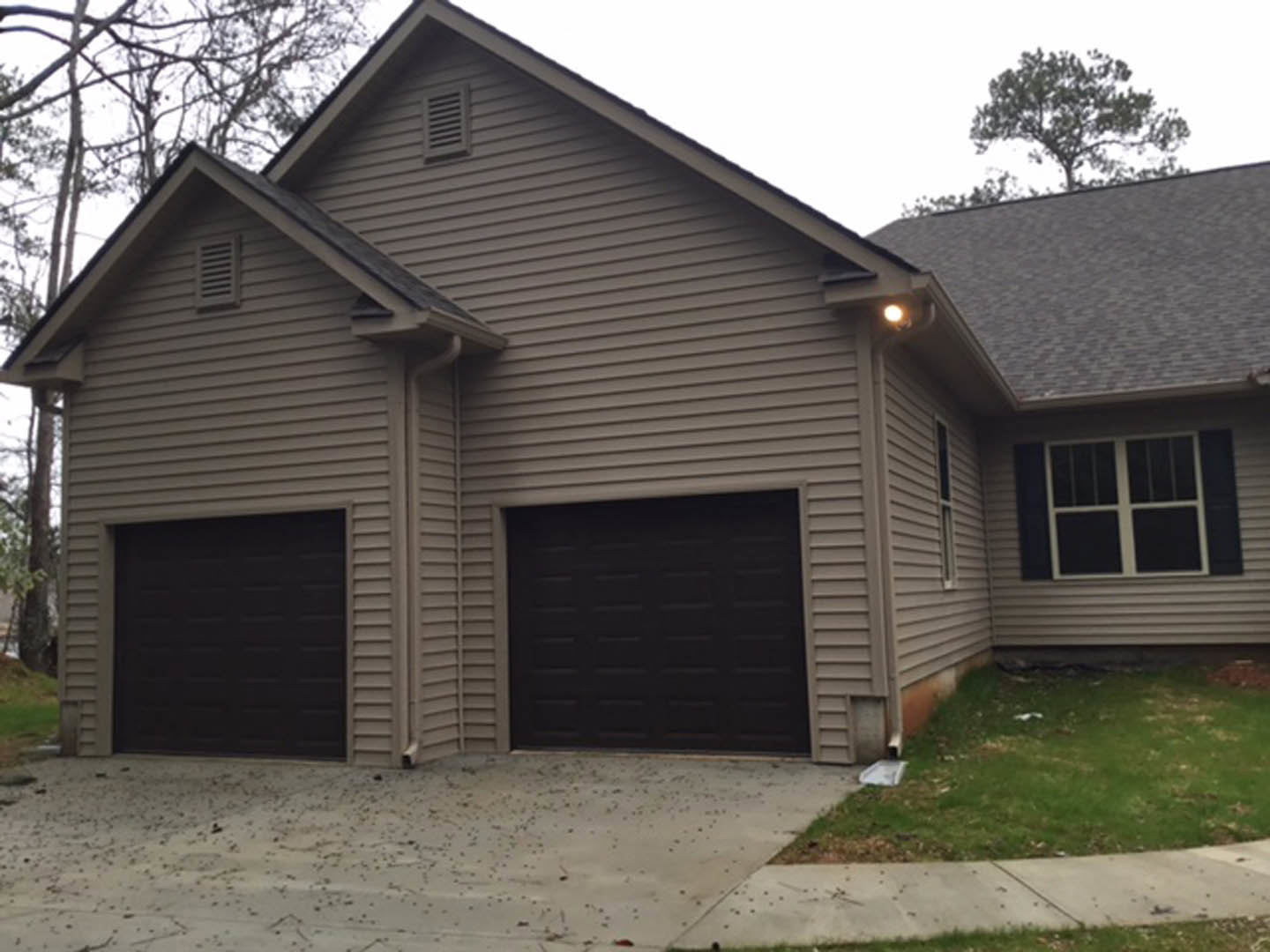 Black garage door with white trim, white-framed window, gray siding, concrete foundation, green grass lawn, roof vent, blue sky, trees in background