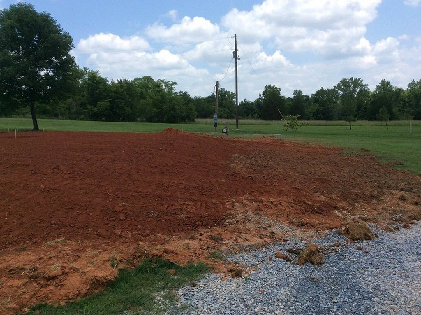 Dirt and gravel field bordered by green-leaved trees, telephone pole with overhead wires, open sky with scattered clouds