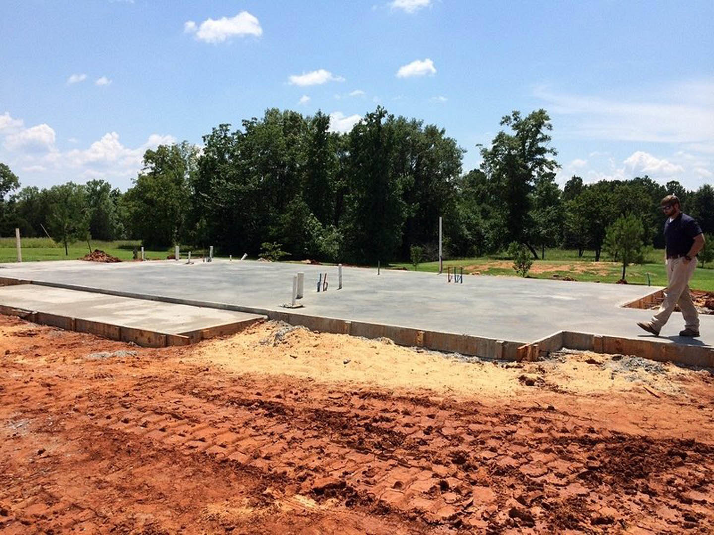 Concrete slab foundation bordered by dirt and a forested area, man standing with hands in pockets, white clouds overhead, dirt road visible near trees