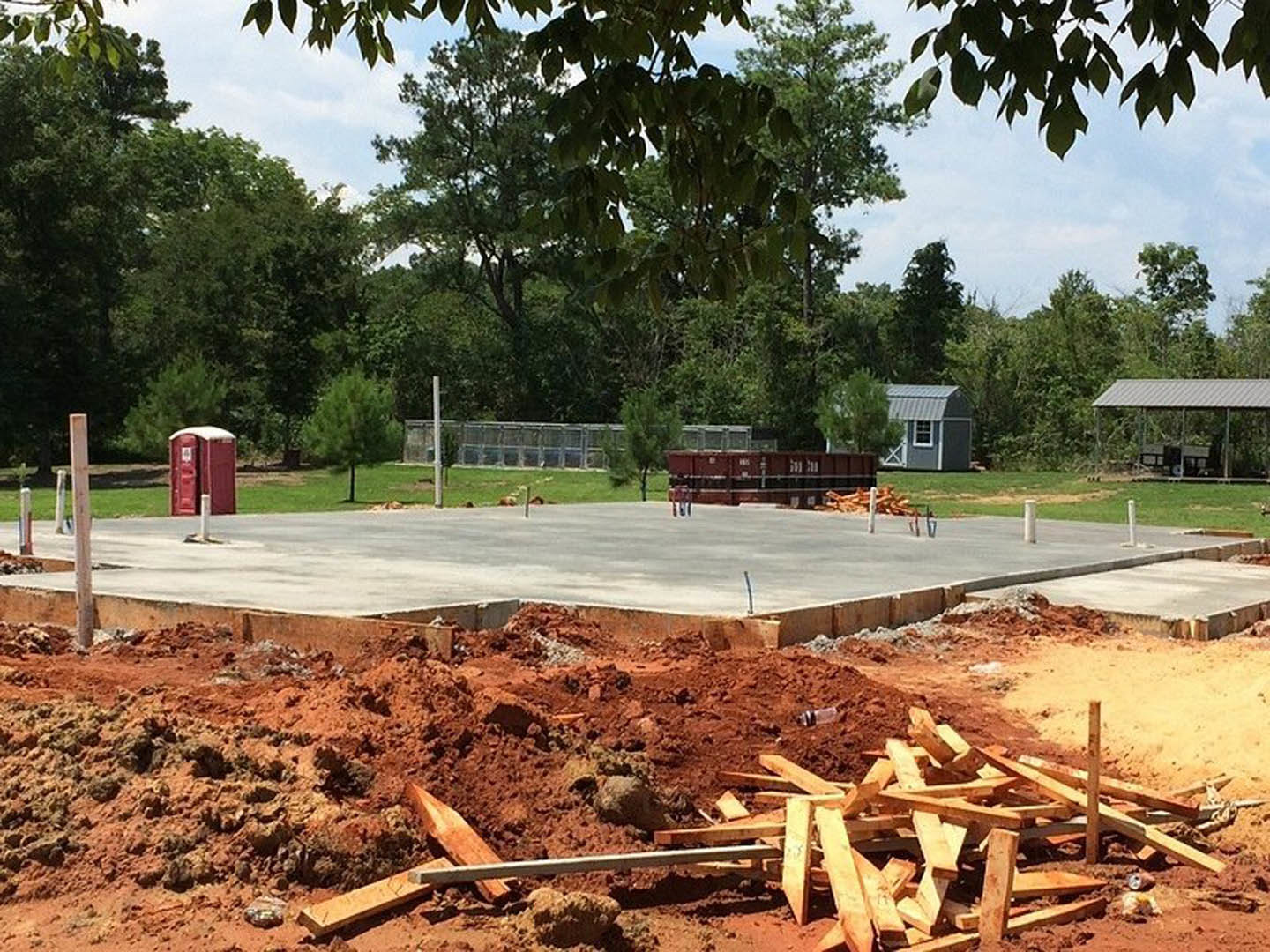 Construction site with stacked lumber, red portable toilet on grass, small shed featuring a white window, metal roof overhang, and trees in the background