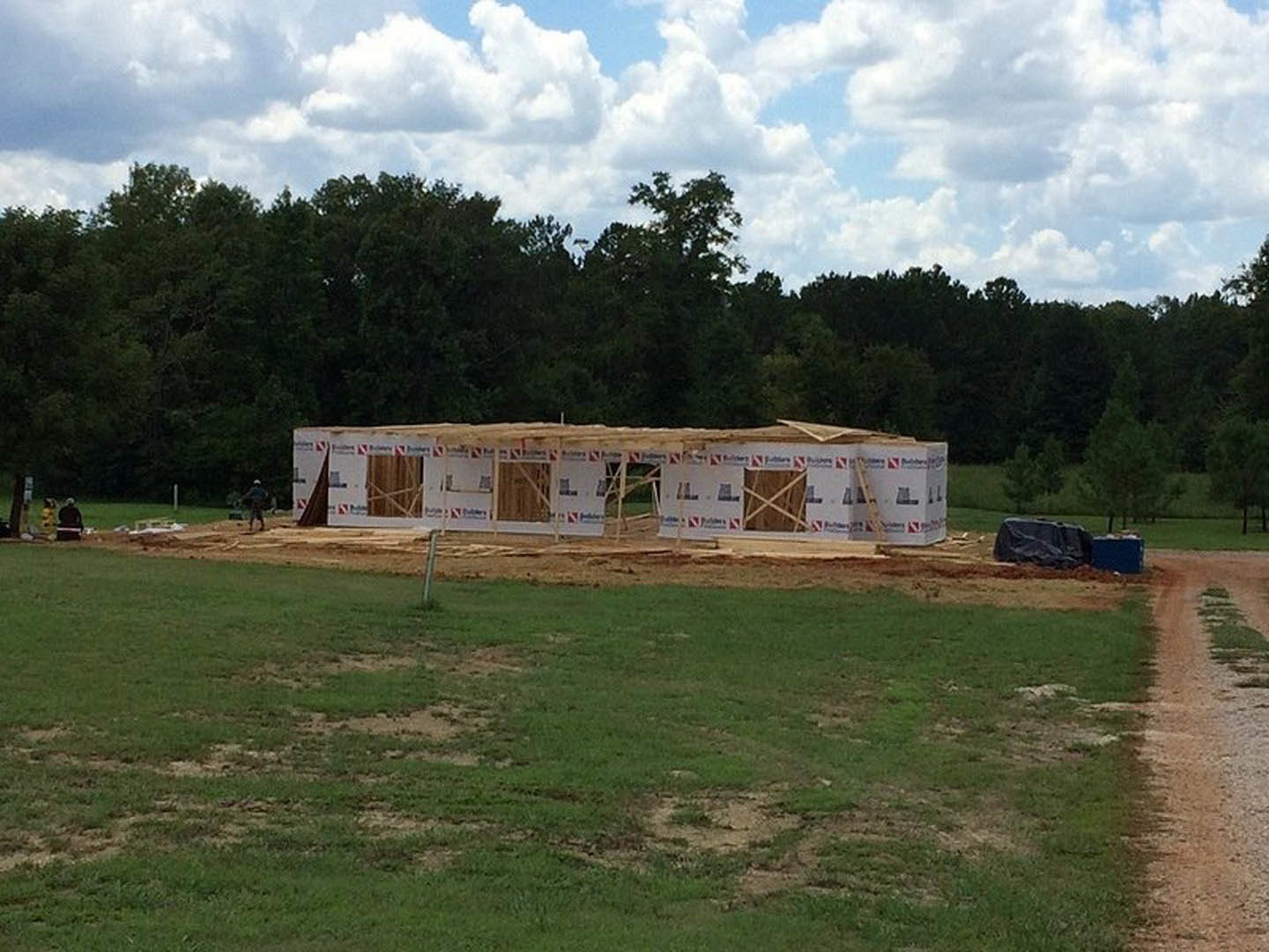 Wood-framed house under construction on a grassy field, exposed plywood walls, broken window, dirt road in foreground, blue sky with scattered clouds, trees lining background