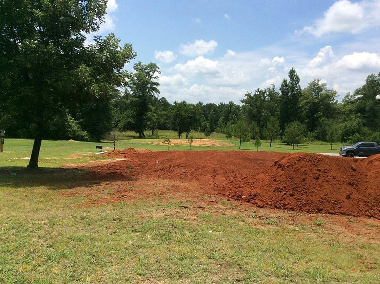 Large mound of soil on grassy field with black pickup truck parked nearby, single tree in background under partly cloudy blue sky