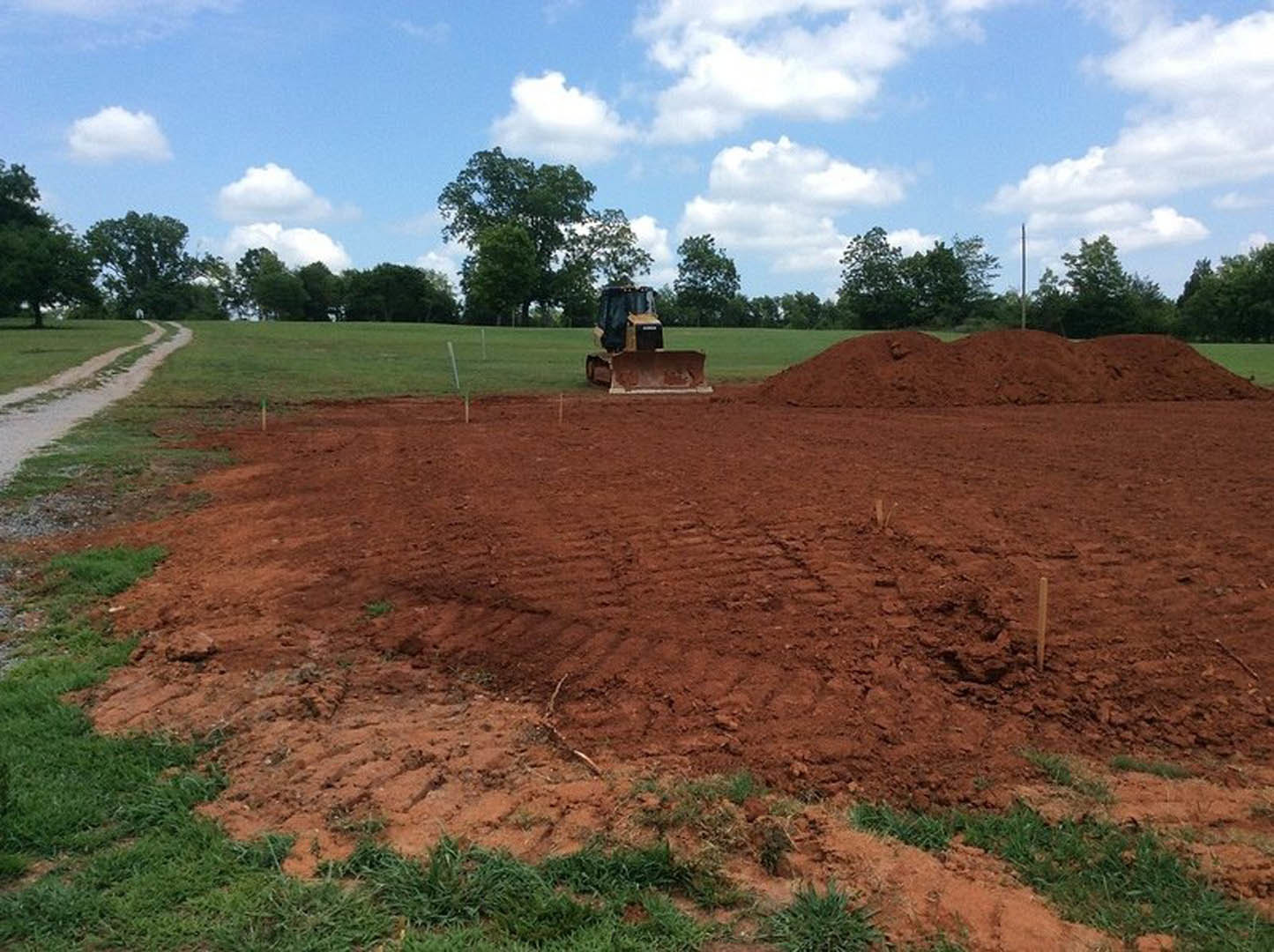 Bulldozer parked on a dirt road in a grassy field, surrounded by trees and under a blue sky with scattered clouds, dirt hill and utility pole visible in the background