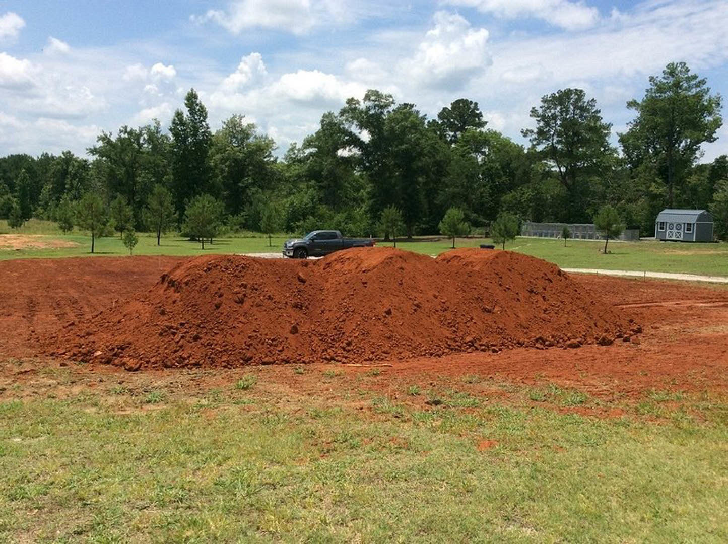 Large mound of soil in grassy field, small grey shed with white trim nearby, black truck parked on street, car in parking lot, blue sky with scattered clouds and trees in