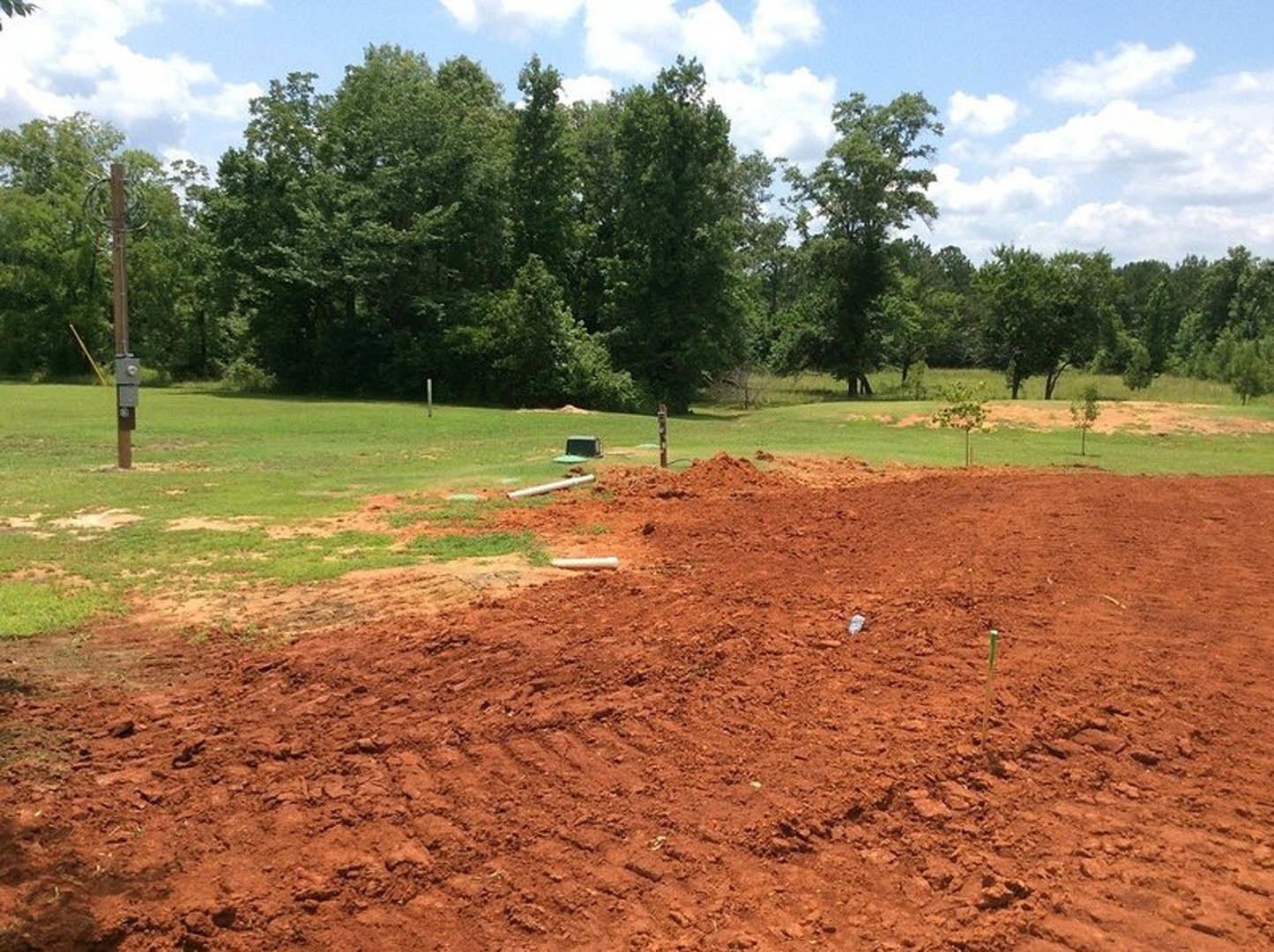 Dirt field bordered by leafy trees under a partly cloudy sky, with patches of grass and a signpost visible in the background