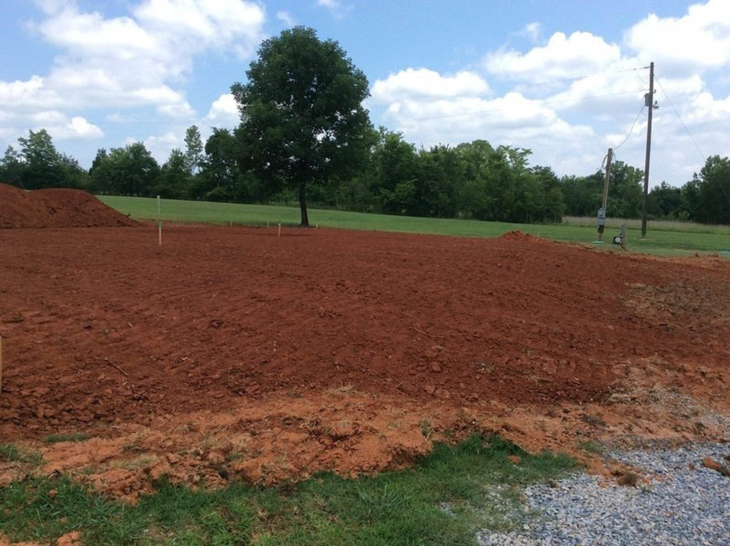 Dirt field bordered by green trees under a blue sky with scattered clouds