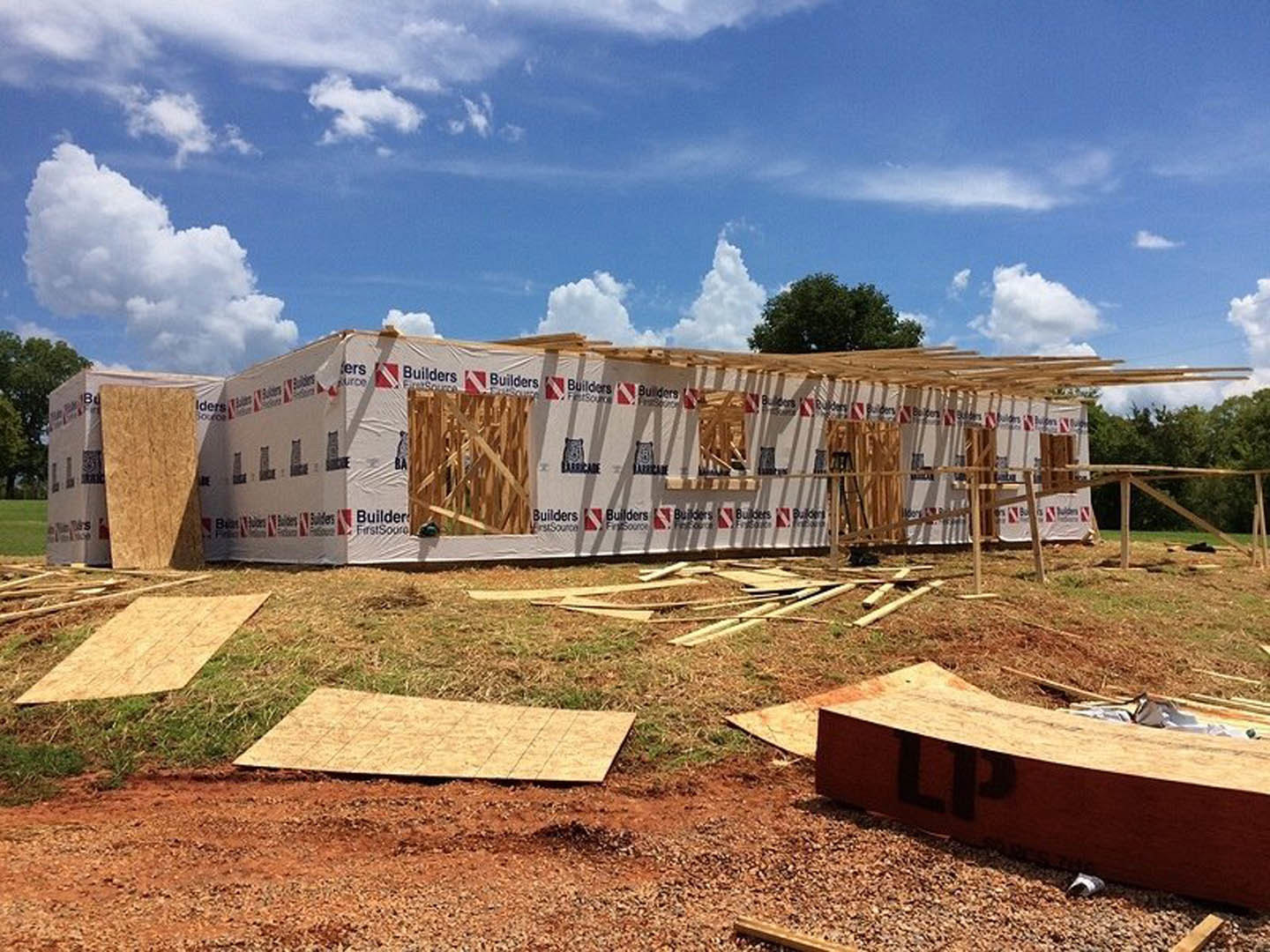 Wood-framed custom home under construction with exposed beams, white exterior wall, pile of lumber on ground, blue sky and tree in background