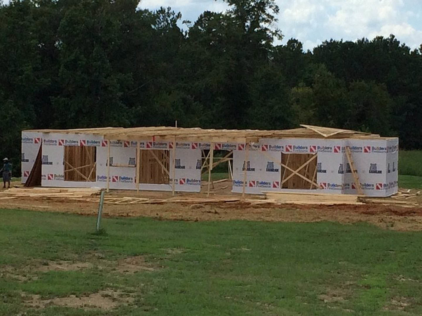 Wood-framed house under construction on a dirt lot, surrounded by grass and mature trees, cloudy sky overhead