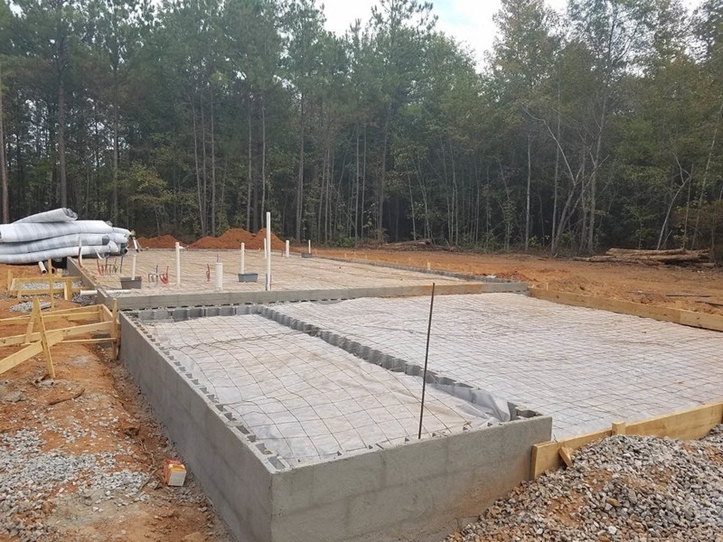 Concrete house foundation with embedded wire mesh, surrounded by bare ground, scattered trees, and a wooden structure nearby under an open sky