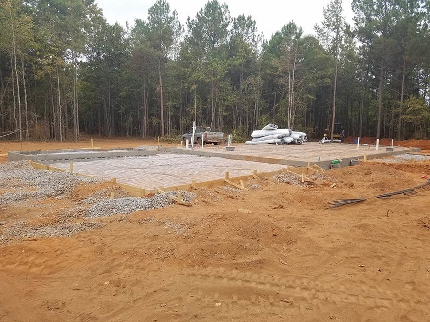 Concrete foundation surrounded by dirt and gravel, grey truck parked nearby, trees in background, construction materials stacked on site