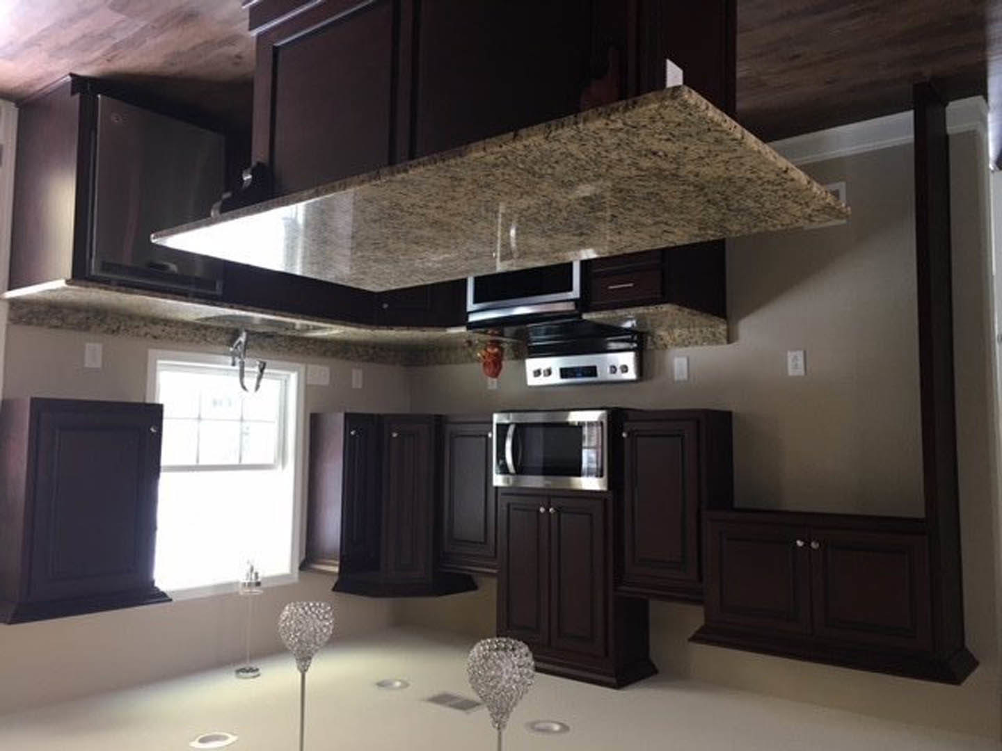 Dark wood kitchen cabinets, white marble countertop, stainless steel microwave, range hood, glass window with sunlight, modern appliances