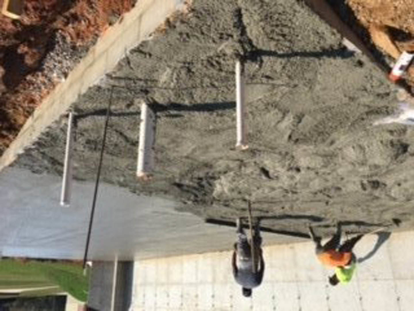 Men in work clothes building a concrete wall outside a house, surrounded by construction materials on bare ground