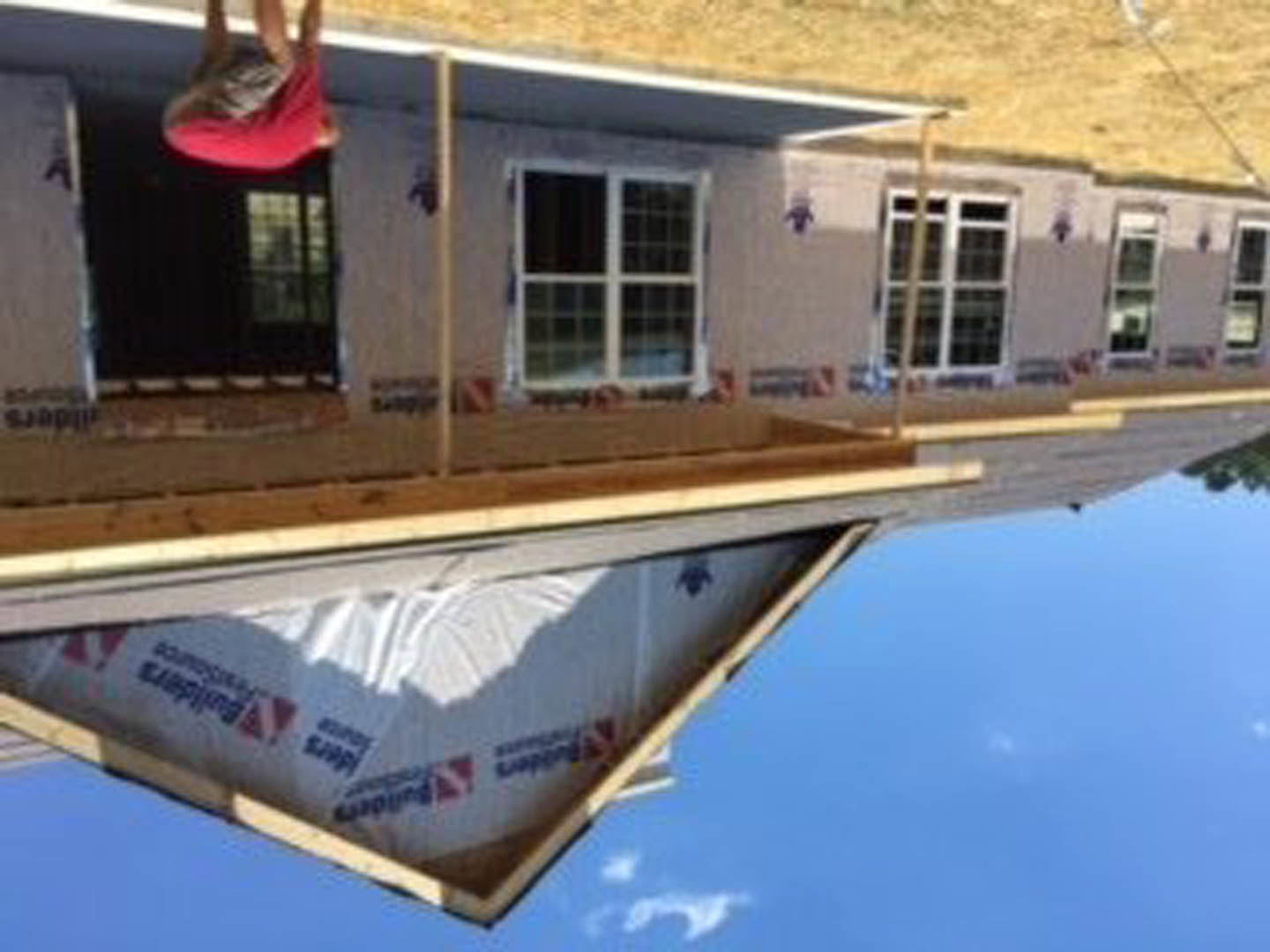 Person in red shirt standing on covered front porch with multi-pane window and shingle roof visible