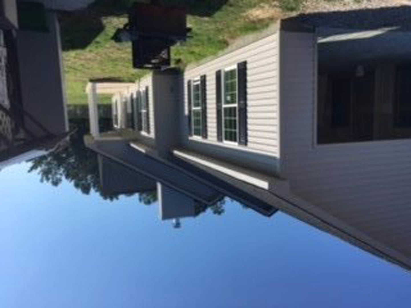 Two-story home with white-framed windows, gray siding, manicured green lawn, mature trees in the background, and clear blue sky
