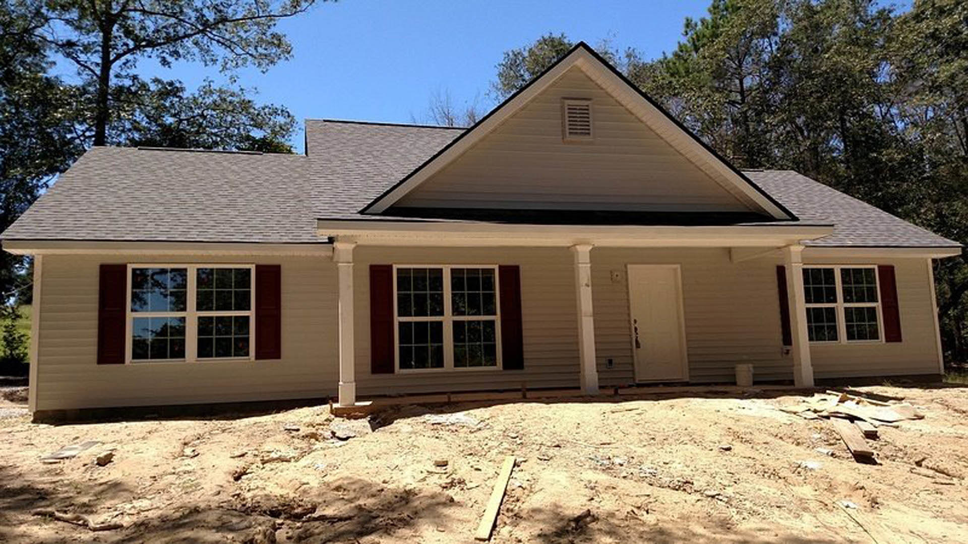 Partially built home with exposed wooden framing, white door installed, vent on exterior wall, large window, scattered wooden planks on ground, surrounded by trees in background