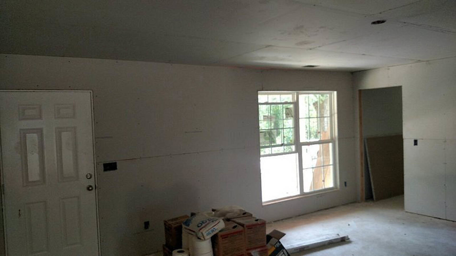 Bright room with plaster walls, large window with clear glass pane, white door with silver doorknob, light-colored flooring, and stacked cardboard boxes near a black rectangular