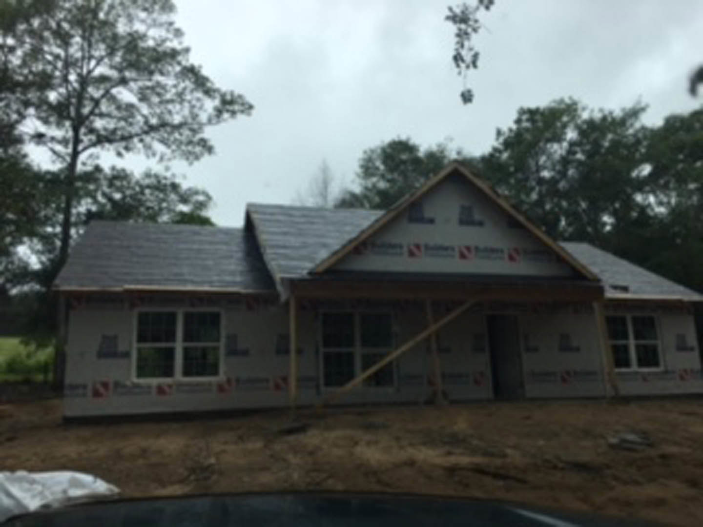Wood-framed house under construction with exposed beams and plywood, surrounded by tall trees and cloudy sky in the background