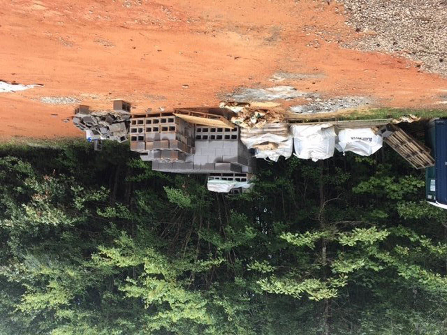 Stack of red bricks and wooden pallets on a dirt driveway beside a building, scattered debris and trash visible, trees and mountain in background