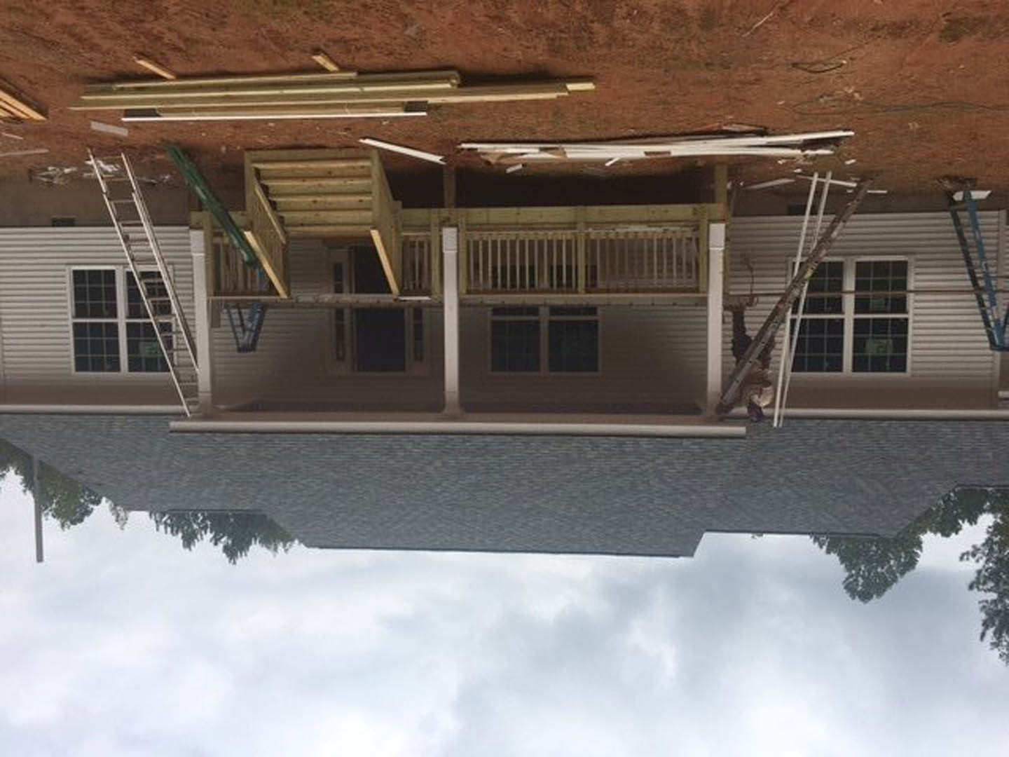 Wood-framed porch with white railing, aluminum ladder leaning against window, partially finished siding, cloudy sky reflected in glass