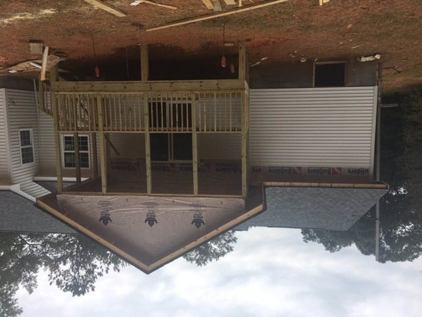 Partially finished home with shingled roof, screened porch, white tarp-wrapped exterior walls, closed garage door, and surrounding trees under cloudy sky