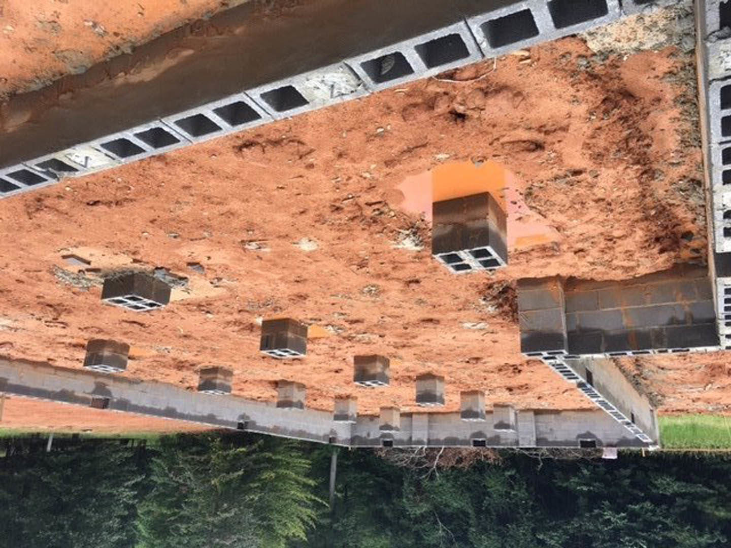Concrete block foundation on dirt construction site, surrounded by trees and plants, with a window opening in a brick wall.