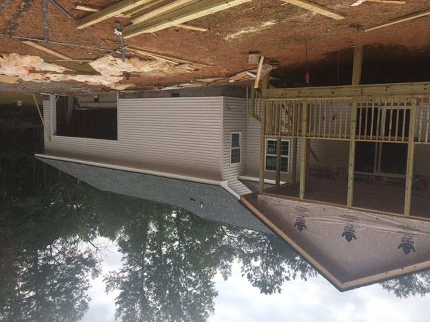 Wood-framed house under construction with exposed wooden deck, white tarp featuring black logo, large windows, and trees reflected in nearby lake
