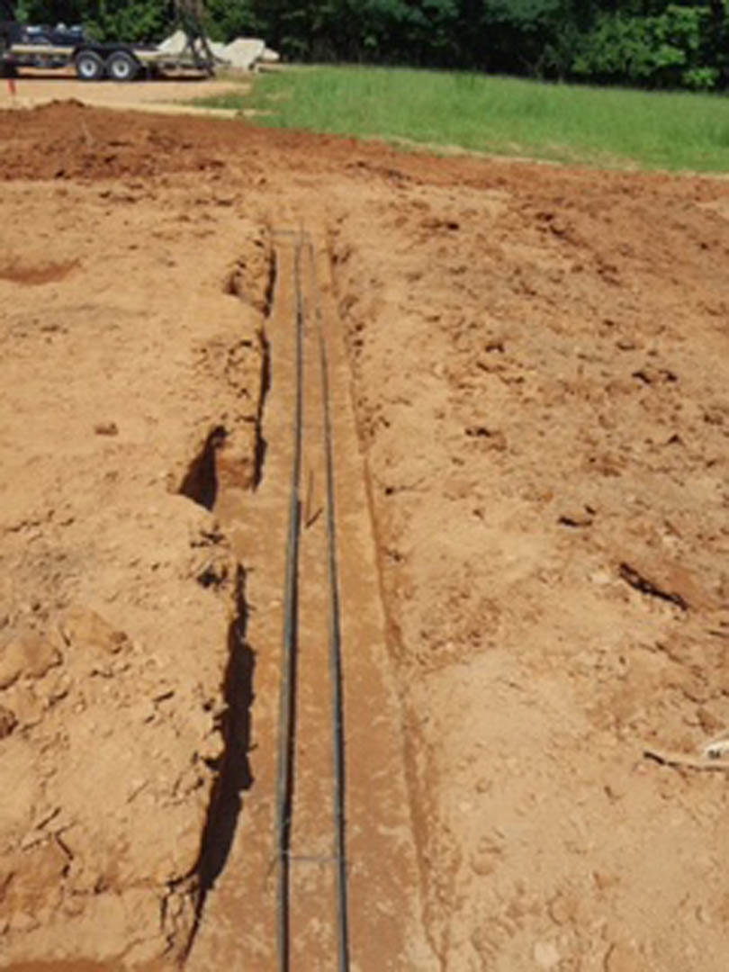Wide view of muddy trench bordered by grass and trees, exposed soil and dirt visible along outdoor construction site