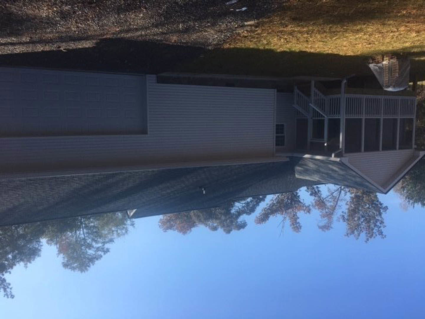 White house with triangular roof, large front porch featuring wooden railings, mature tree in landscaped yard, blue sky and lake reflected in windows