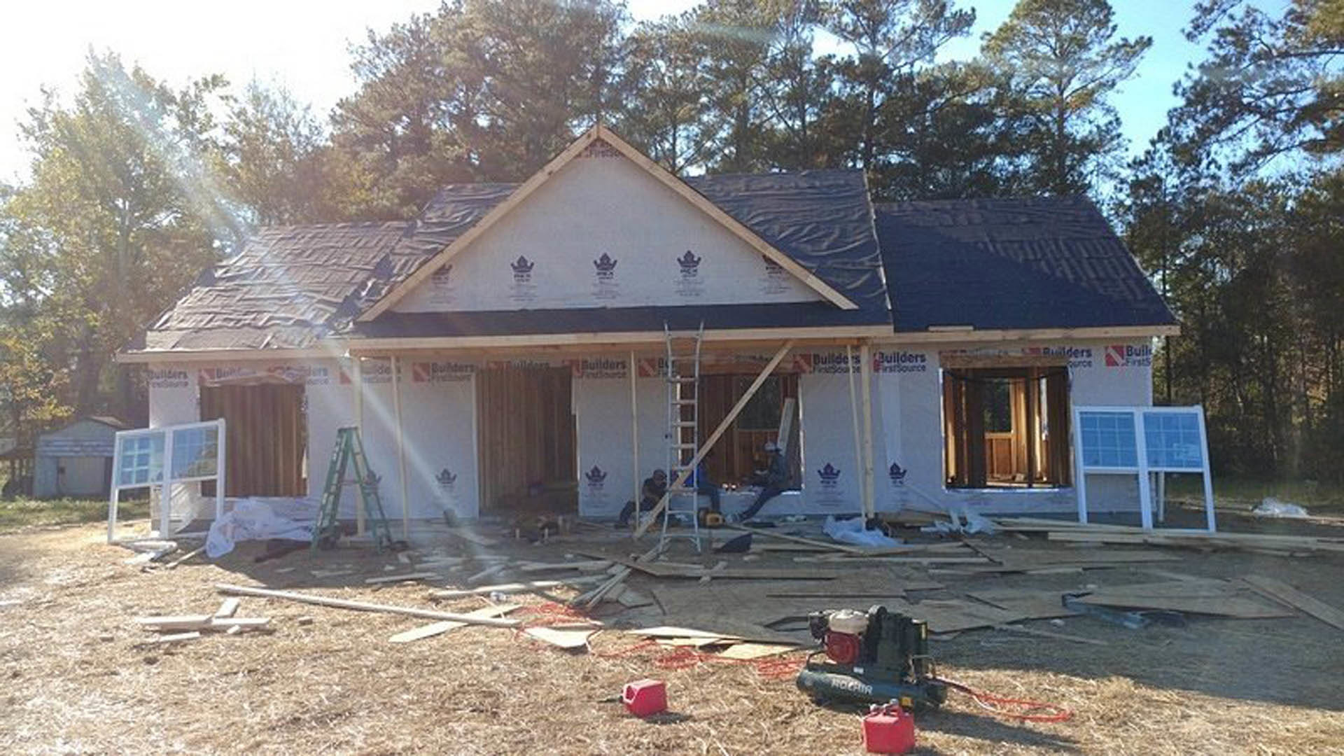 Partially built house with exposed wooden framing, two ladders leaning against exterior wall, window frame installed, scattered lumber and construction debris on ground