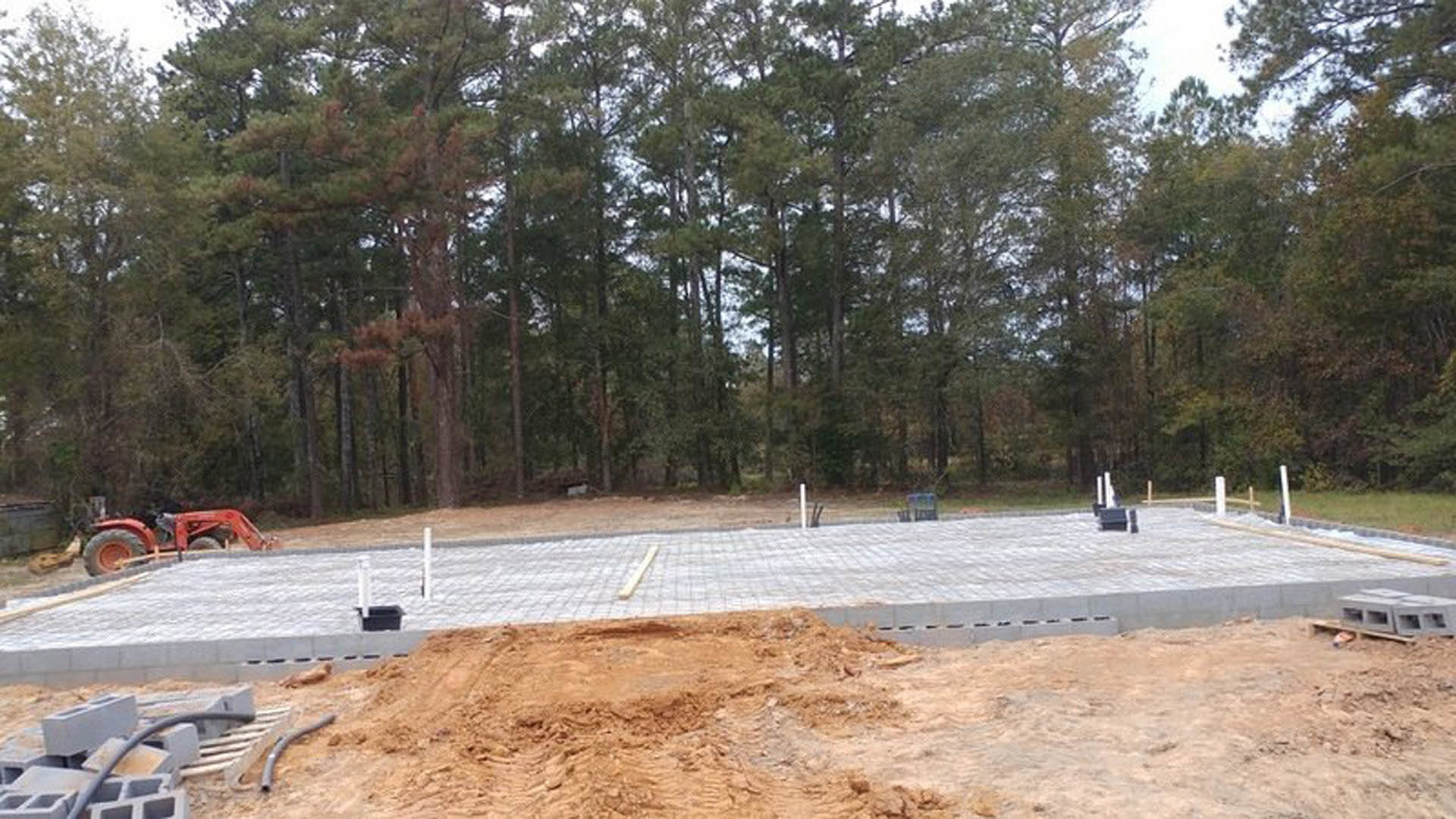 Concrete foundation slab with wood beam, red tractor parked on snowy dirt, forested trees in background